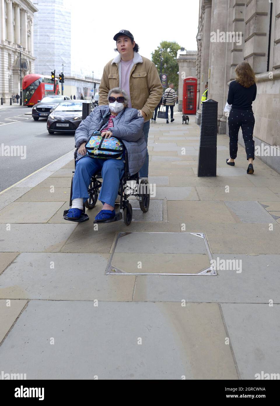 London, England, Großbritannien. Mann, der eine Frau mit Gesichtsbefrag in einen Rollstuhl schiebt, 2021 Stockfoto