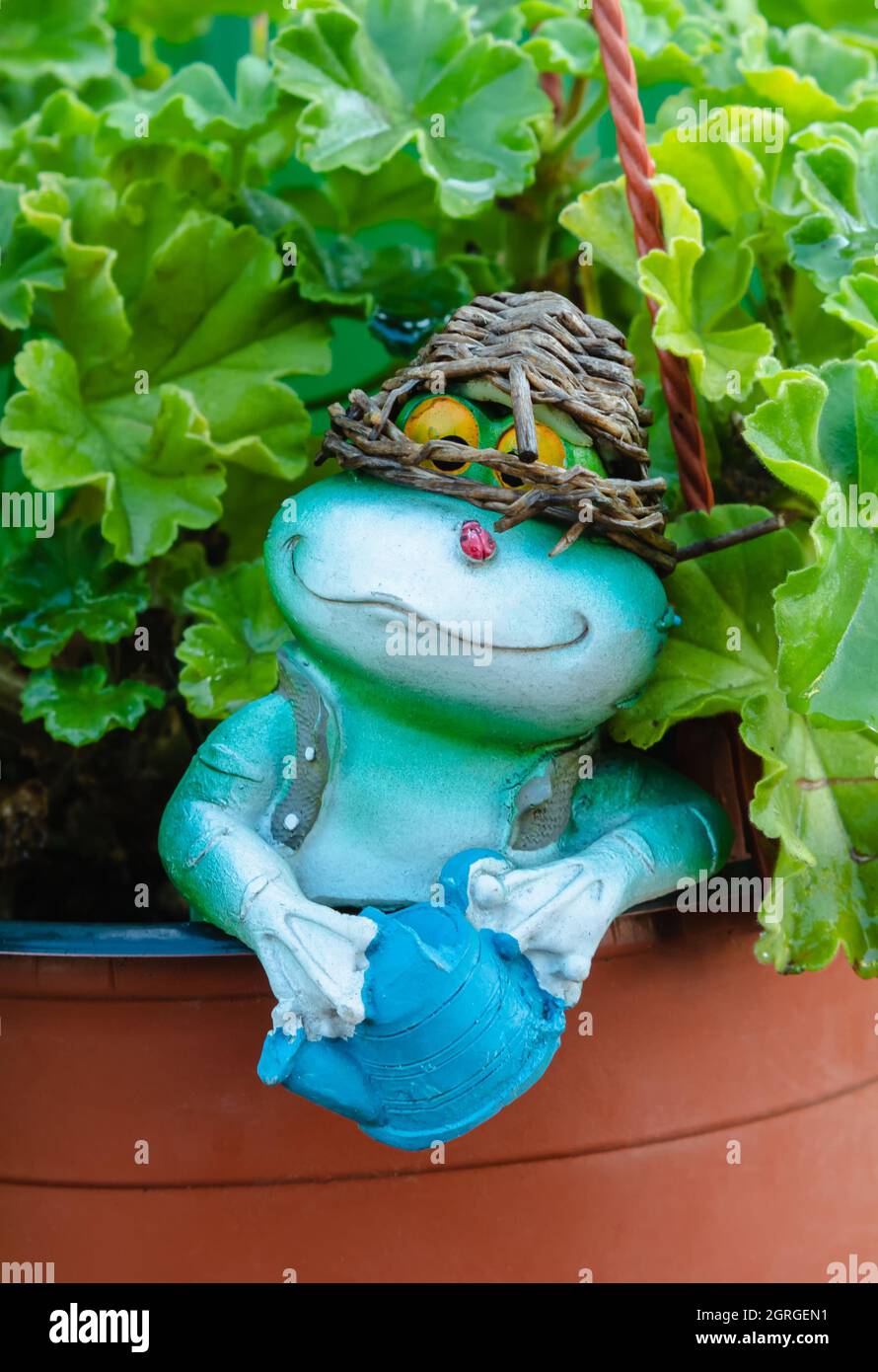 Gartenfigur eines Frosches mit einem Korb auf dem Kopf, zwischen grünen Blättern. Dekoratives Spielzeug, das in einem Topf mit einer Blume steht Stockfoto