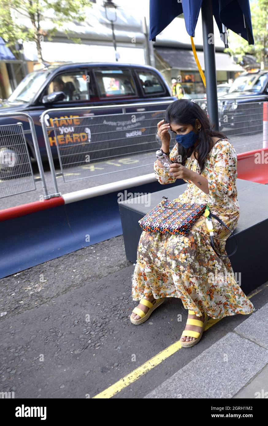 London, England, Großbritannien. Junge Frau in einem blumigen Kleid mit COVID-Maske und Mobiltelefon Stockfoto
