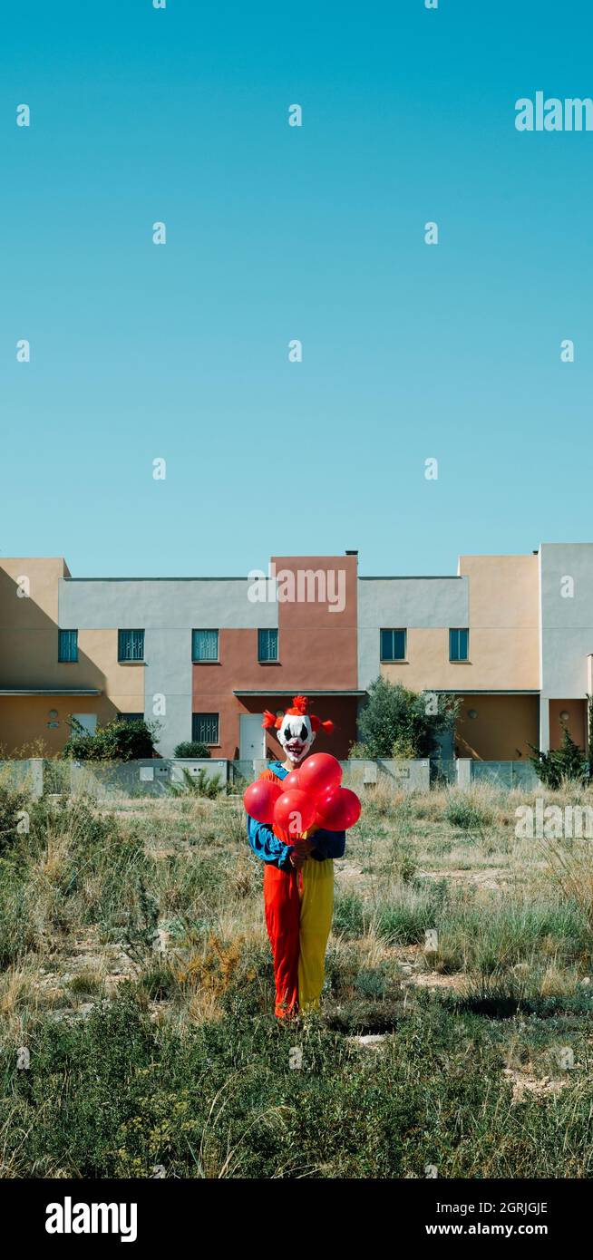 Ein gruseliger Clown, der ein farbenfrohes Kostüm trägt, einen Haufen roter Ballons in der Hand hält, auf einem leeren Grundstück steht, in einem vertikalen Format, das für den Mob verwendet werden kann Stockfoto