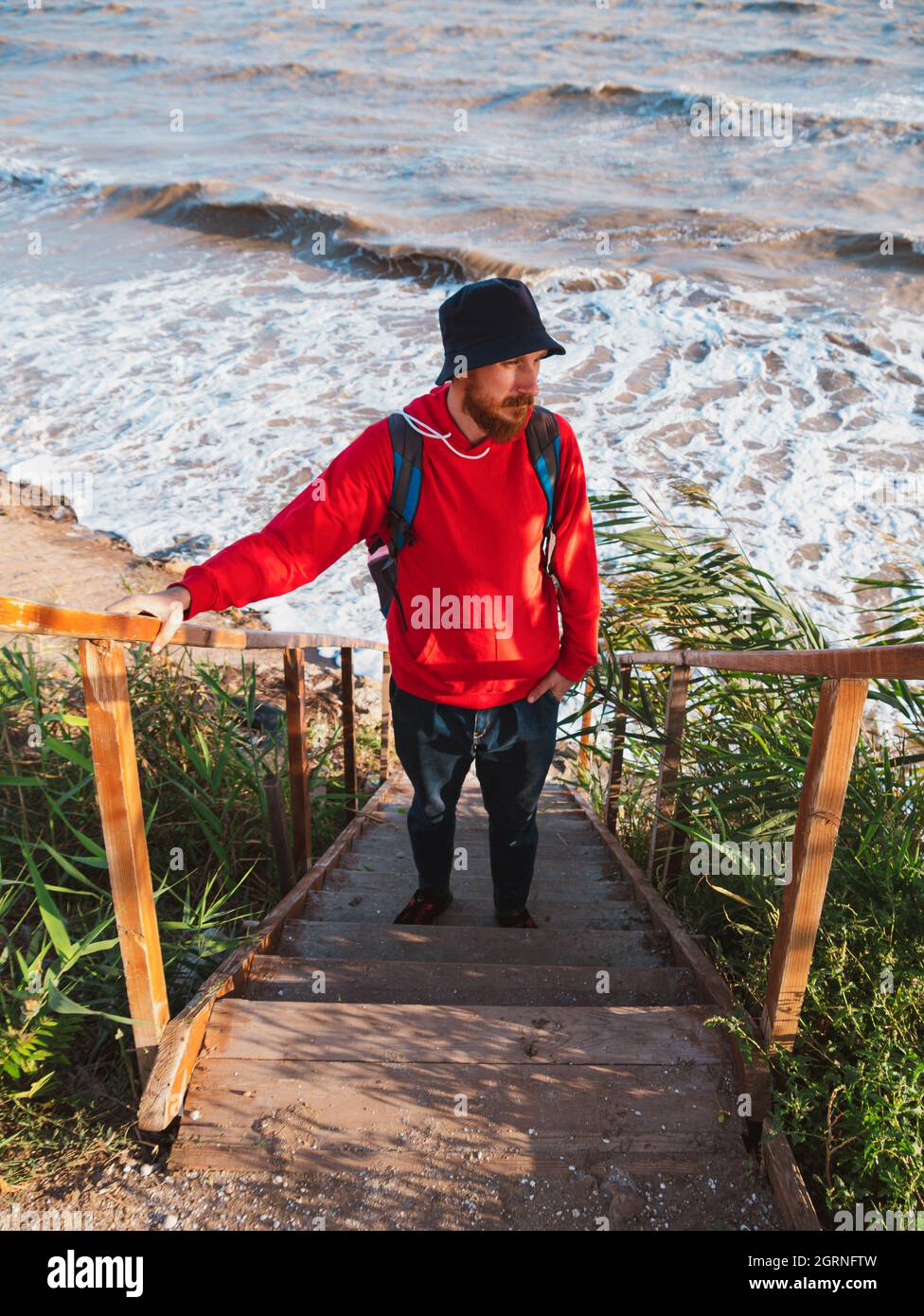 Bärtiger Tausendjähriger Mann in rotem Kapuzenpullover mit Rucksack Herbst Meer Hintergrund trockene Schilf authentische männliche Tourist Lifestyle Foto Stockfoto