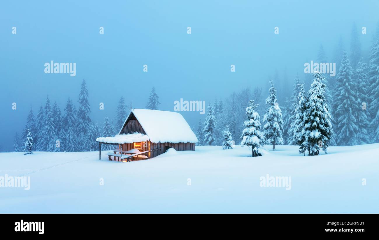 Fantastische Winterlandschaft mit glühender Holzhütte im verschneiten Wald. Gemütliches Haus in den Karpaten. Weihnachtsfeiertagskonzept Stockfoto