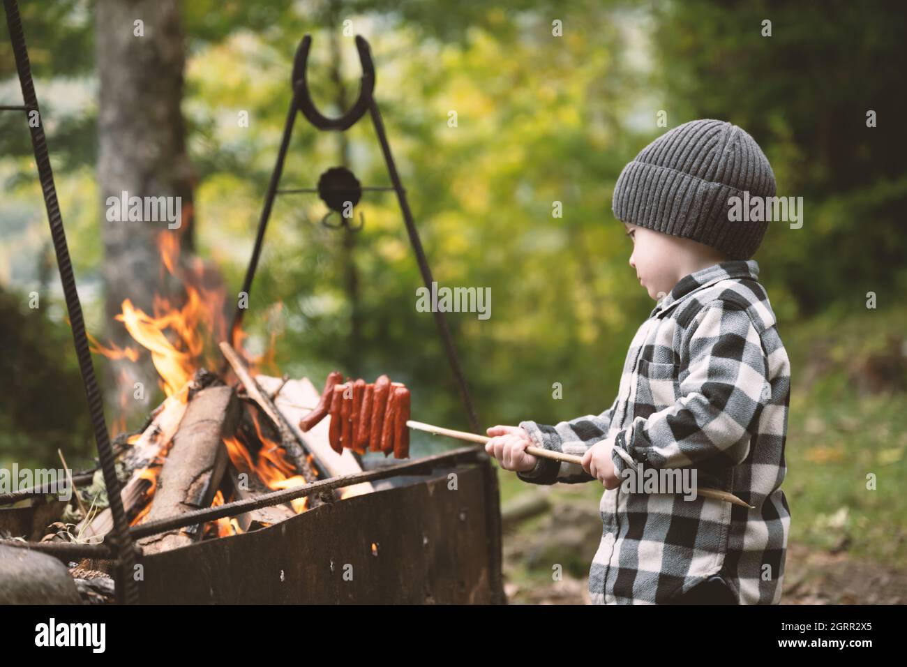 Ein kleines Kind in einem karierten Hemd und einem grauen Hut fritiert Würstchen am Lagerfeuer im Wald. Kindheit mit naturliebenden Konzept Stockfoto
