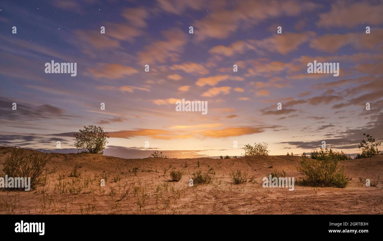 Leuchtende Wolken und sandige Landschaft mit Sternenhimmel bei Sonnenaufgang. Astronomische Landschaft mit langer Belichtung Stockfoto