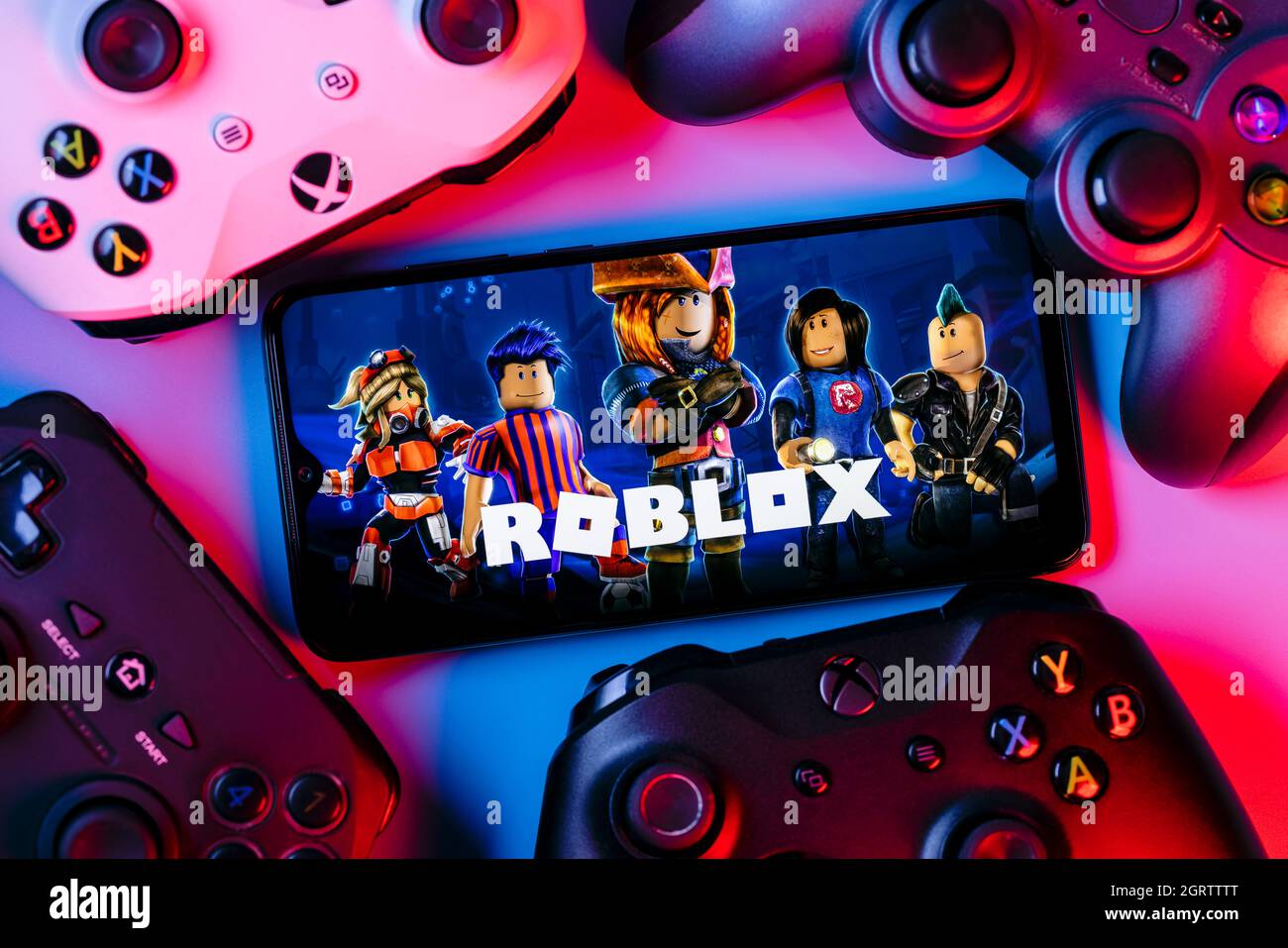 Roblox ist eine Online-Spielplattform und ein System zur Erstellung von Spielen. Ein Smartphone mit dem Roblox-Logo auf dem Bildschirm, umgeben von Gamepads. Stockfoto