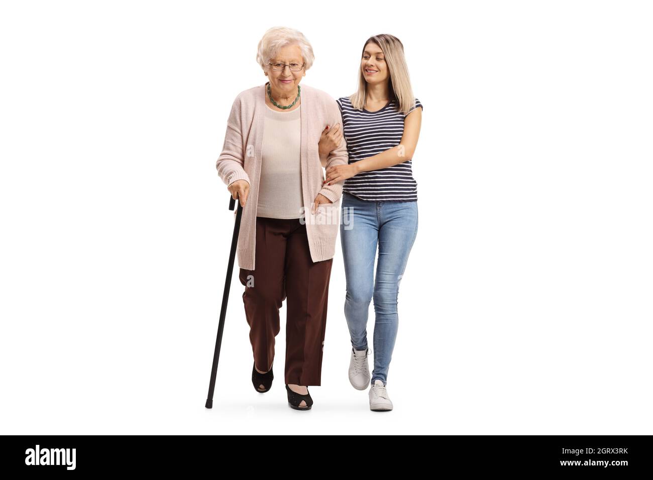 In voller Länge Porträt einer freundlichen jungen Frau, die einer älteren Frau hilft, die mit einem Gehstock auf weißem Hintergrund isoliert geht Stockfoto