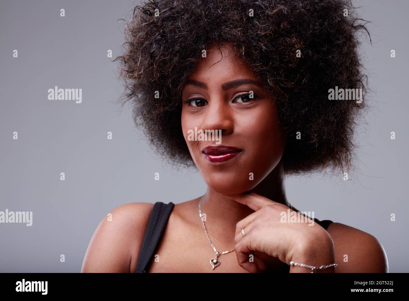 Süße, junge schwarze Frau mit einem unruhigen Grinsen und ihrem Finger bis zum Kinn lächelt leise auf die Kamera in einem Nahaufnahme-Porträt über einem grauen Studio Stockfoto