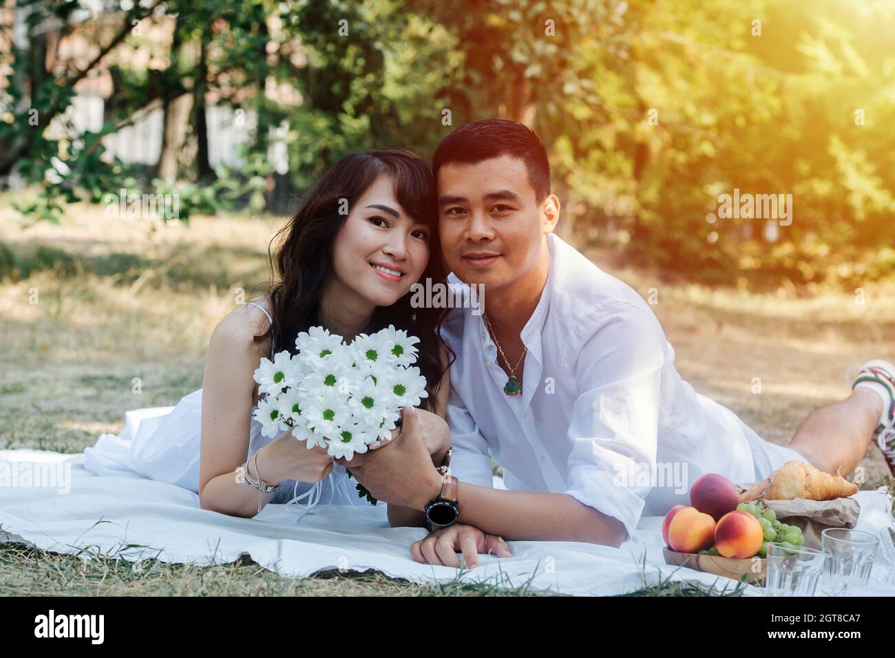 Schönes asiatisches Paar happing Picknick in einem Park, liegen auf ihren Mägen. Frau mit Strauß, die in weißen Kleidern in einem Baumschatten ruht. Stockfoto