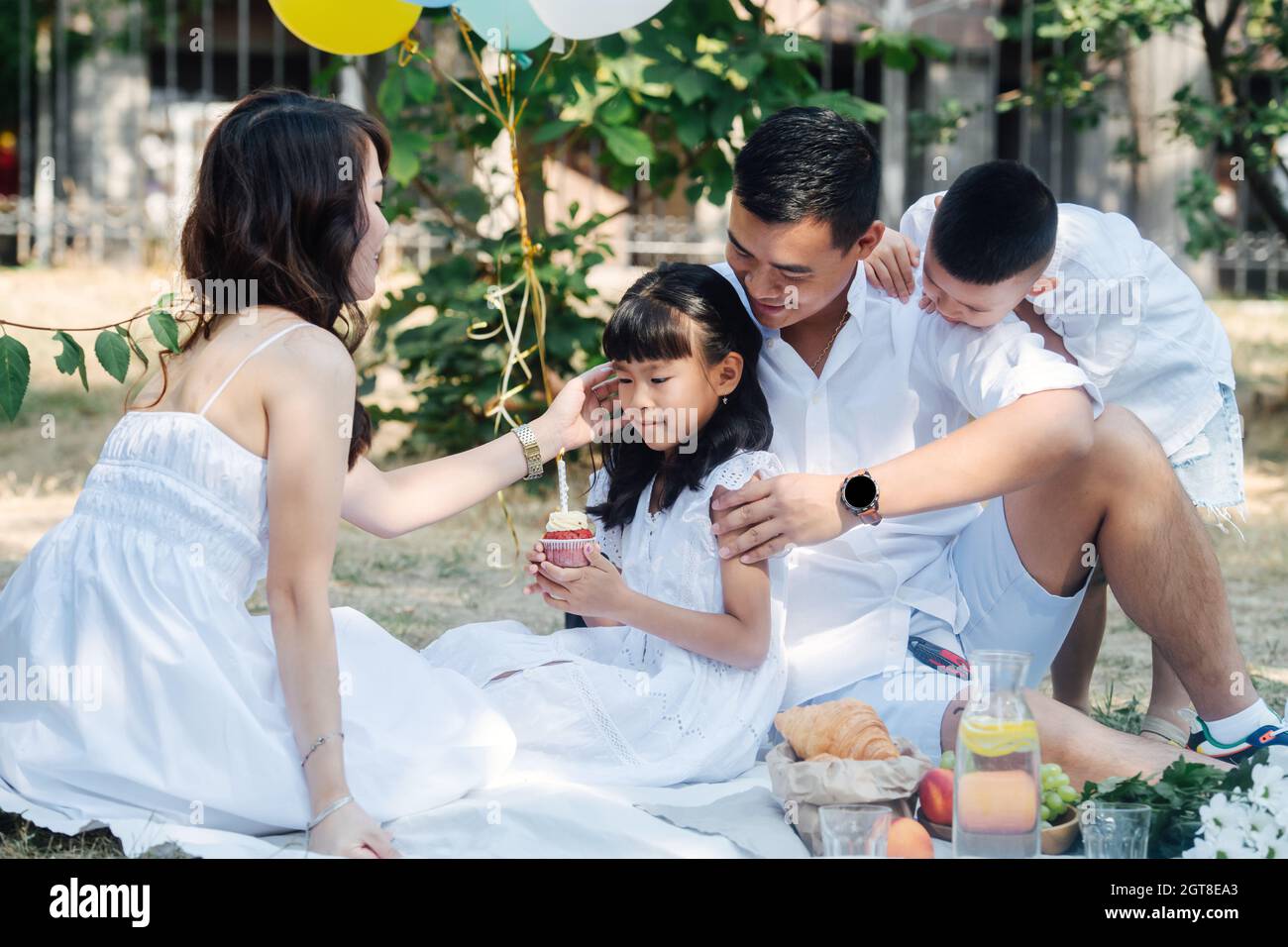 Fürsorgliche asiatische Familie feiert den Geburtstag ihres kleinen Mädchens in einem Park mit einem kleinen Kuchen. Eltern und ihre Kinder in weißen Kleidern im Baumschatten Stockfoto