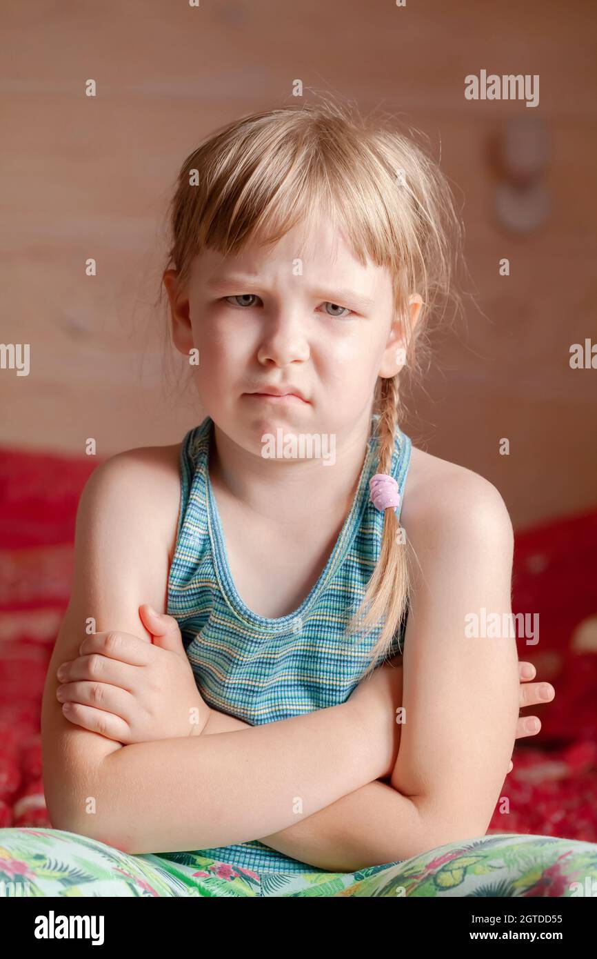 Ein einziges beleidigtes Kind, kleines Mädchen im Schulalter, das zu Hause mit gekreuzten Armen auf dem Bett sitzt, unglücklich, rebellisches Kind einfaches vertikales Porträt, Stockfoto
