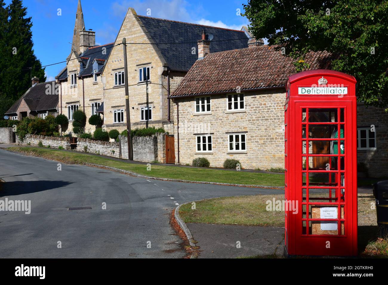 Eine alte rote Telefonbox, die für einen Defibrillator umfunktioniert wurde, neben dem Village Green in Barnwell, Northamptonshire, Großbritannien Stockfoto