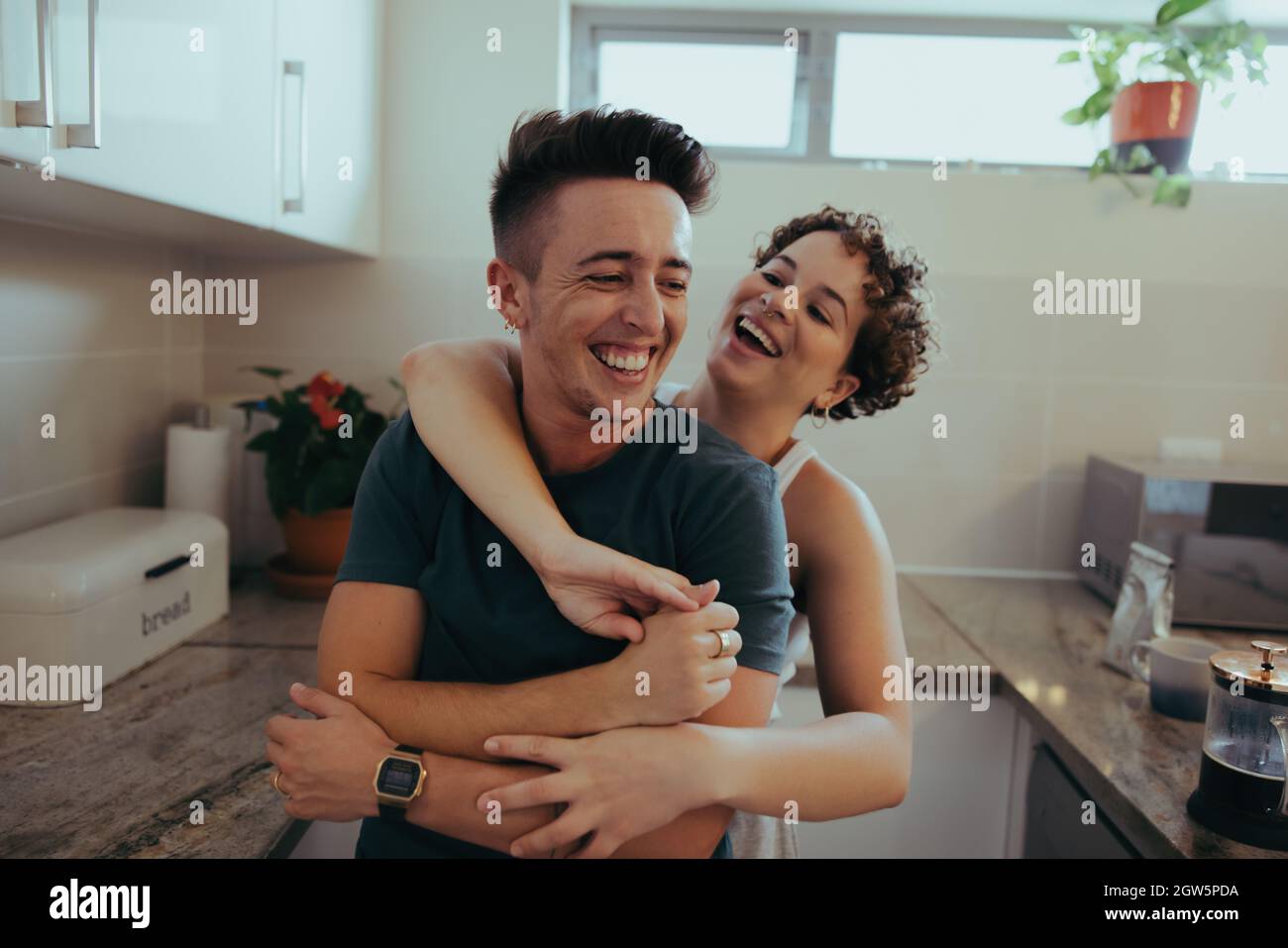 Romantisches queeres Paar umarmt sich gegenseitig und lacht zusammen in ihrer Küche. Glückliches junges queeres Paar, das am Morgen Spaß zusammen hat. Jung Stockfoto
