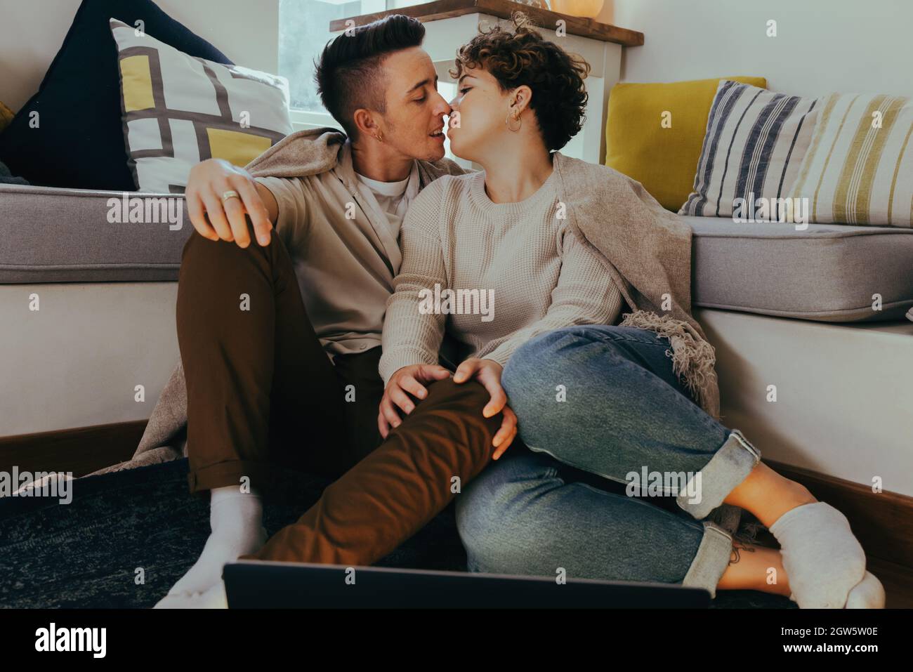 Süßes queeres Paar, das im Wohnzimmer einen Kuss macht. Romantisches junges Paar, das sich küsst, während es im Wohnzimmer zusammen sitzt. Young L Stockfoto