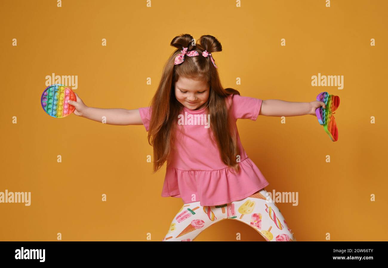 Frolic Kind Mädchen in bunten Kleidern hält zwei sensorische Regenbogenfarben Spielzeug - Pop it und schaut nach unten. Spielen und Tanzen Stockfoto