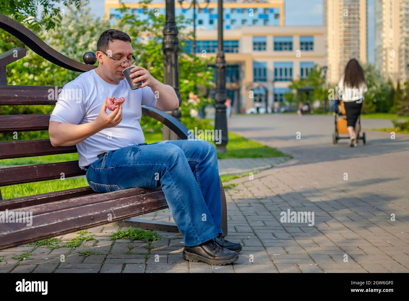 Der Blick auf einen Mann, der lässig Kaffee, Tee, Kakao mit einem Brötchen und Donut trinkt und auf seine Hose in einem Stadtpark auf einer Bank tropft Stockfoto