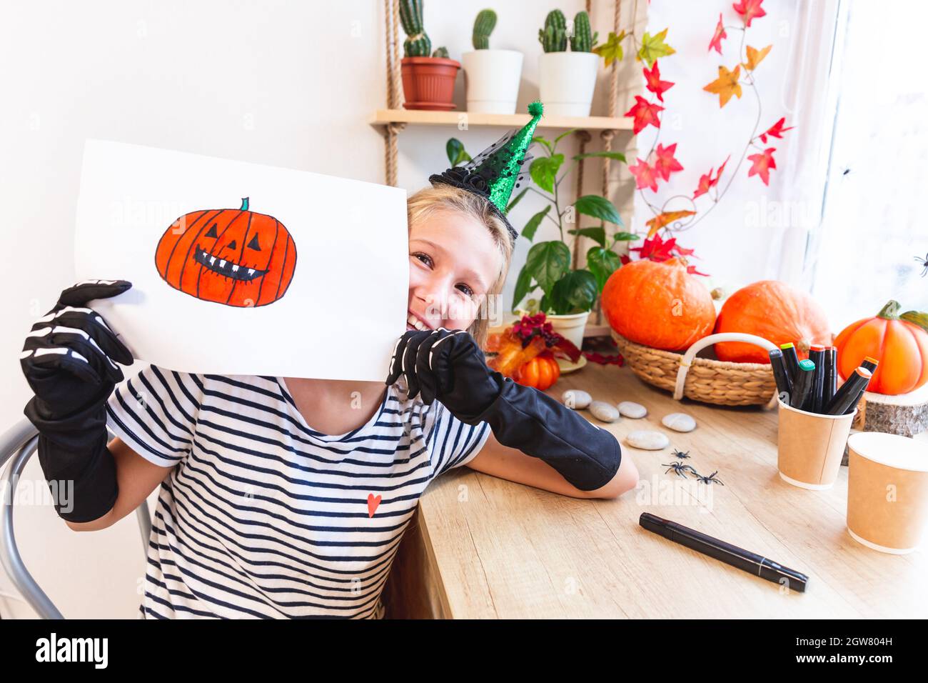 Ein fröhliches Mädchen in einem Hexenkostüm zeigt ihre Zeichnung an Halloween, von hinten heraus blickend, an einem Tisch am Fenster sitzend, umgeben von Stockfoto