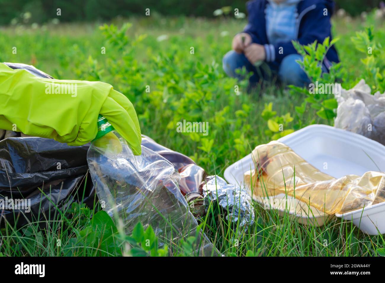 Weibliche Hände in gelben Gummihandschuhen sammeln Plastikmüll in einem schwarzen Beutel im Freien. Im Hintergrund hilft ein Junge. Selektiver Fokus. Sommer sonnig Stockfoto