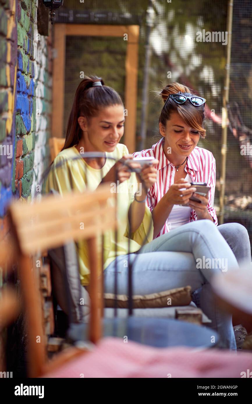 Schöne junge Erwachsene weibliche Freunde sitzen nebeneinander im Café im Freien, Blick auf ihre Handys. Social issue Konzept Stockfoto