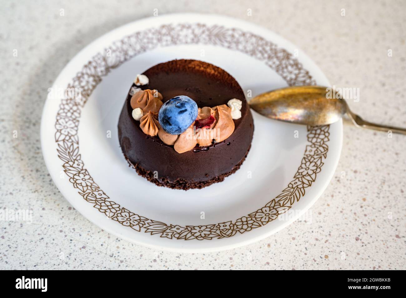 Abgerundetes Schokoladendessert (Fondant) mit Heidelbeere und Creme auf dekorativem Teller mit goldenem Löffel, Nahaufnahme. Stockfoto