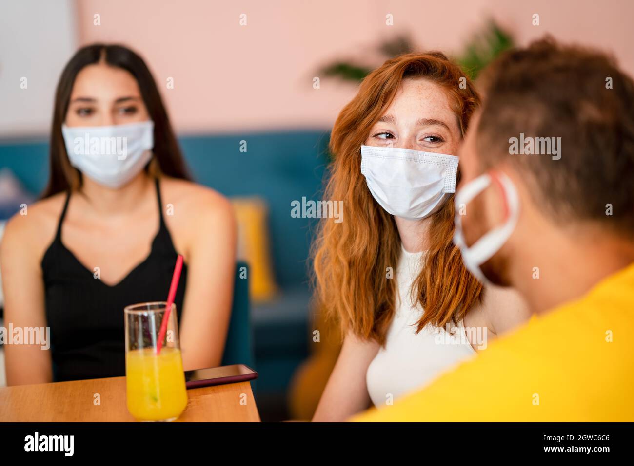 Grops von Freunden plaudern und lächeln, während sie in einem Café mit medizinischer Gesichtsmaske sitzen und ein Getränk trinken. Der Corona-Virus bricht aus. Stockfoto