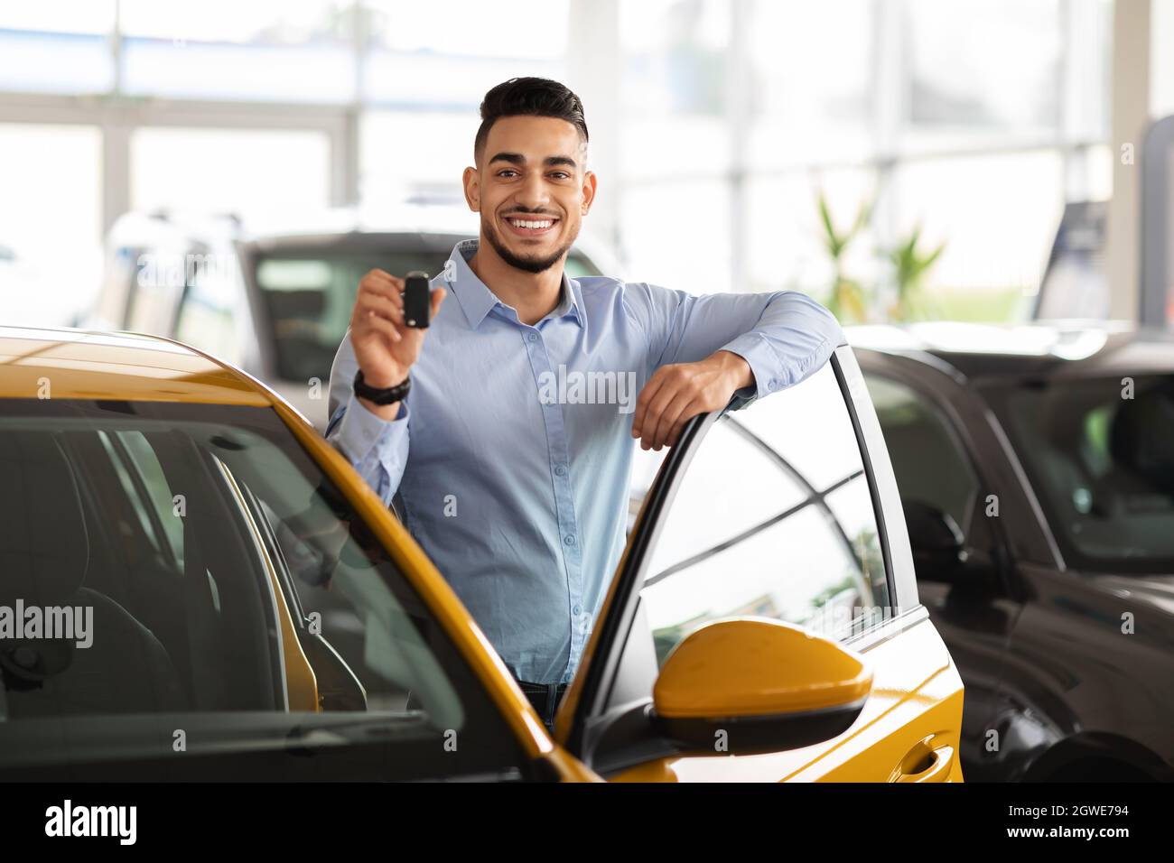 Wohlhabender mittelöstlicher Geschäftsmann, der in einem luxuriösen Autosalon ein neues Auto kauft, fröhlicher arabischer Mann, der neben einem schönen, bequemen gelben Auto steht und Schlüssel zeigt Stockfoto
