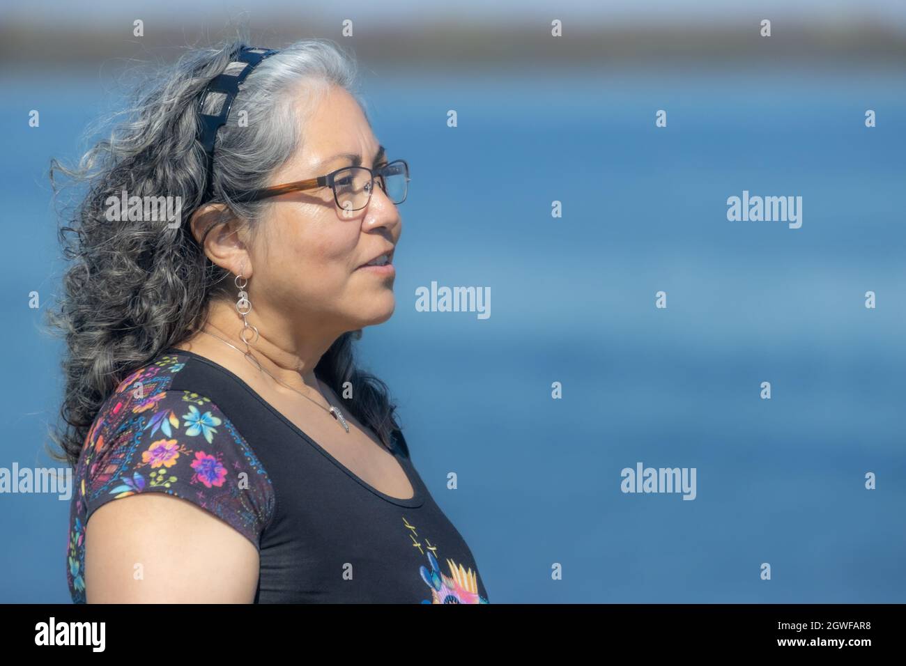 Profilportrait einer nachdenklichen und reflektierenden reifen Latina-Frau, blauer Hintergrund, gebräunte Haut, langes, welliges gräuliches Haar, Stirnband, Brille, kein Makeug Stockfoto