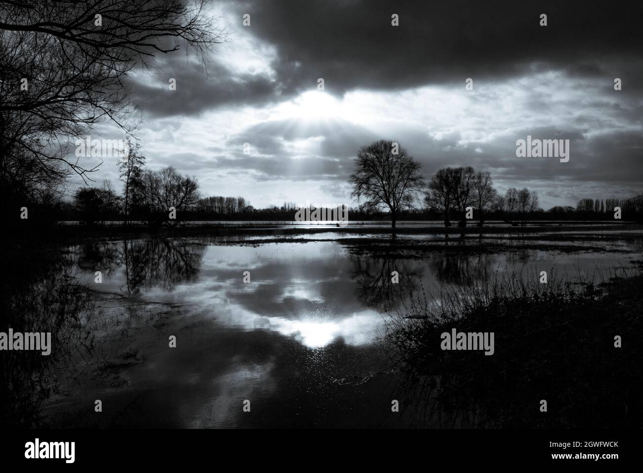 Eine friedliche, ruhige Landschaft in Schwarz und Weiß - die Sonne, die durch Wolken auf das stille, überflutete Wasser des Flusses Great Ouse bricht Stockfoto