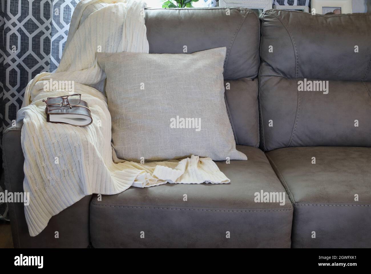 Weiche Strickdecke über den Arm einer grauen Couch in einem Wohnzimmer im Bauernstil mit geschlossenem Buch und Lesebrille. Stockfoto