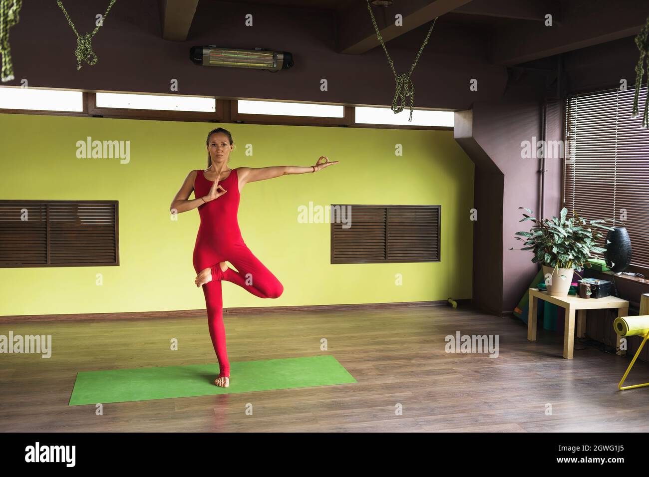 Eine Frau führt Vrikshasana-Übung mit einer Geste des Bewusstseins, Baumhaltung, steht auf einer Matte im Studio, trainiert in einem roten Sport-Jumpsuit Stockfoto