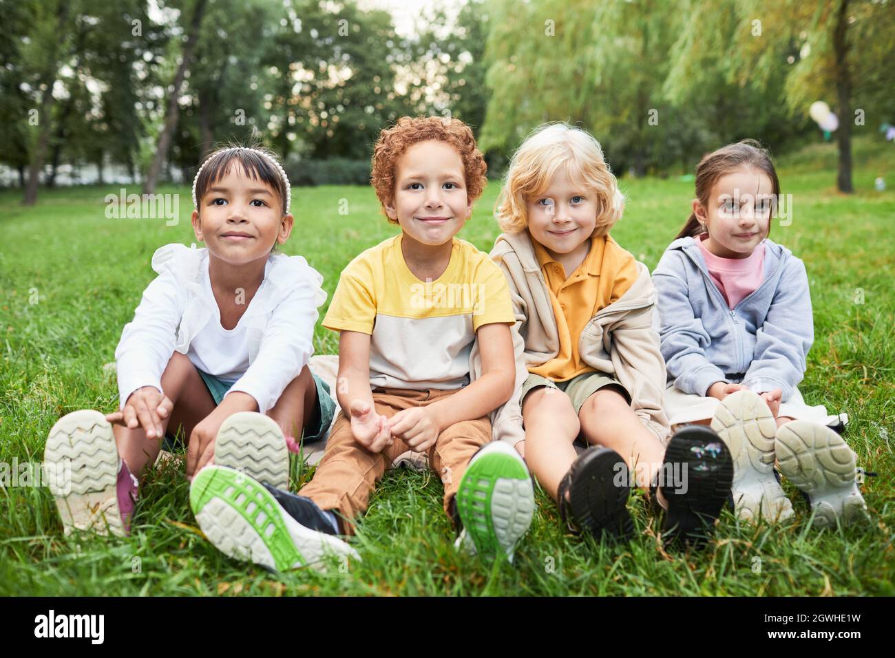 In voller Länge Porträt einer Gruppe von Kindern, die im Park auf Gras sitzen und die Kamera betrachten Stockfoto