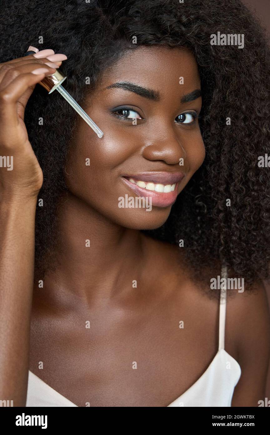 Gepflegtes glückliches schwarzes Mädchen, das pipettierendes Gesichtsserumöl anwendet. Hautpflegekonzept. Stockfoto