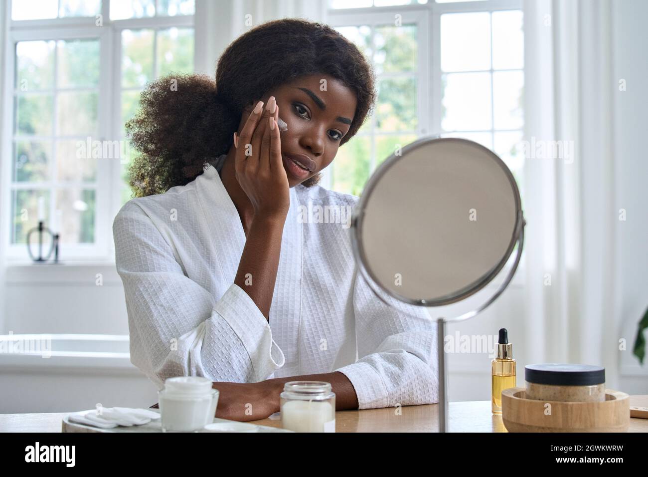 Junge glückliche schwarze Mädchen Anwendung Gesichtscreme vor dem Spiegel im Badezimmer. Stockfoto