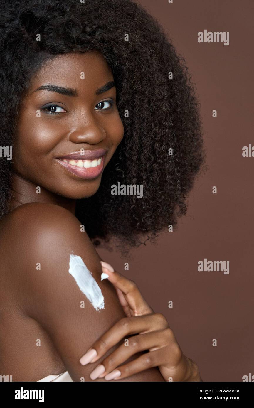Lächelndes, fröhliches junges afro-amerikanisches Mädchen, das Creme auf den Körper aufgibt. Hautpflegekonzept. Stockfoto