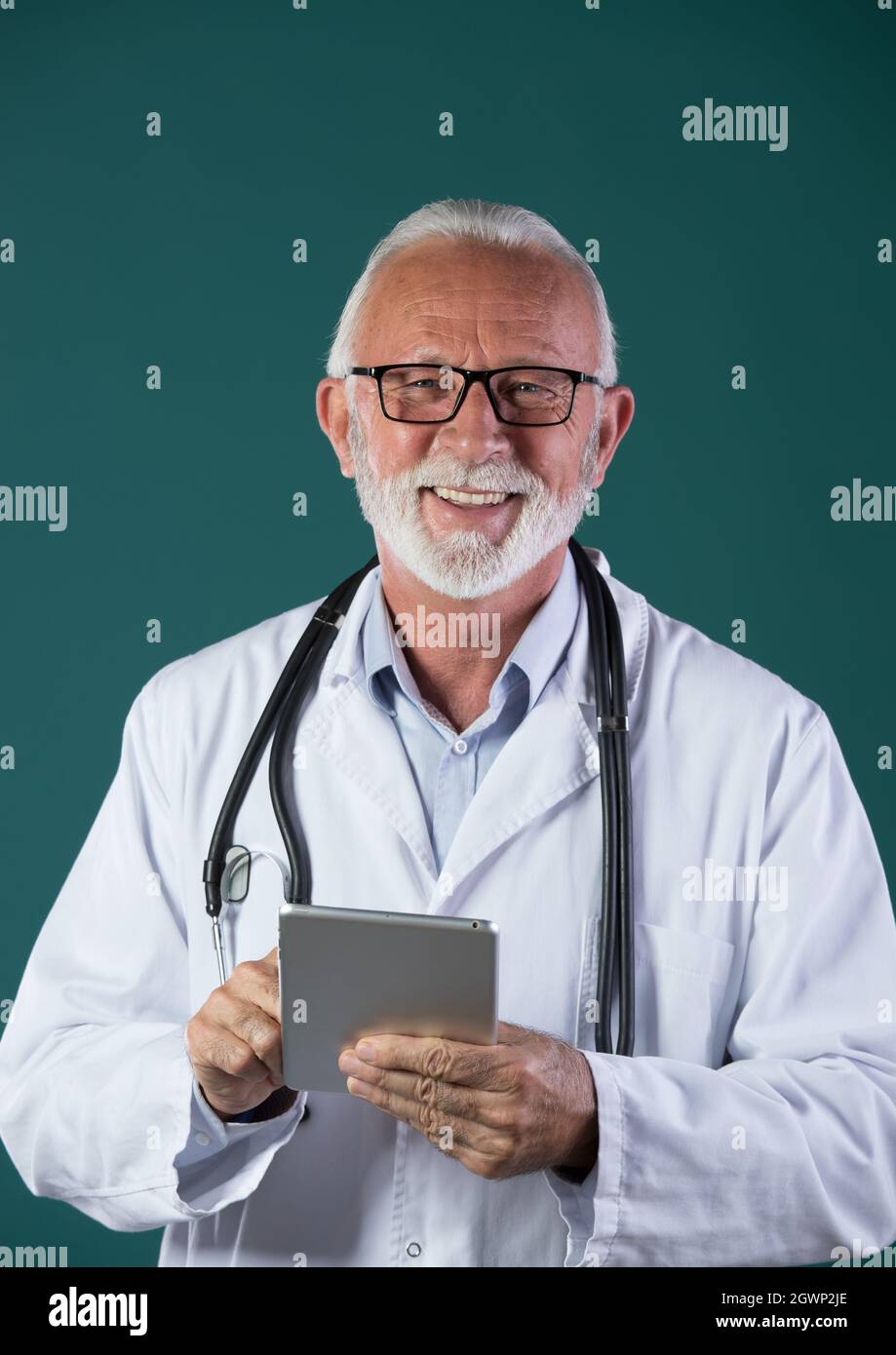 Porträt eines leitenden Arztes mit weißem Mantel, Stethoskop um den Hals, Tablet-PC haltend, lächelnd und vor blauem Hintergrund auf die Kamera schauend Stockfoto
