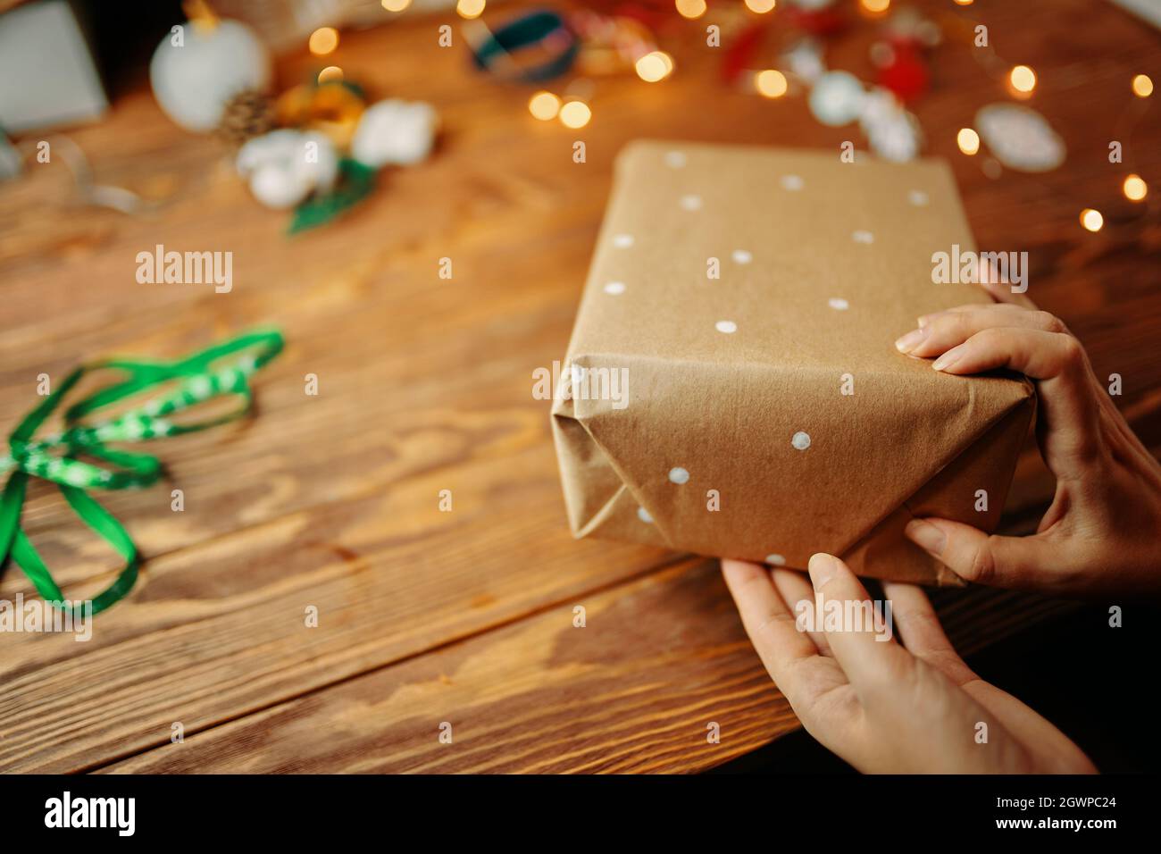 Nahaufnahme von Frauenhänden, die Weihnachtsgeschenke auf einem Holztisch verpacken. Geschenkpapier mit weißen Punkten. Farbbänder Licht Girlande und Neujahr Spielzeug auf dem Hintergrund. Festliche Atmosphäre. Stockfoto