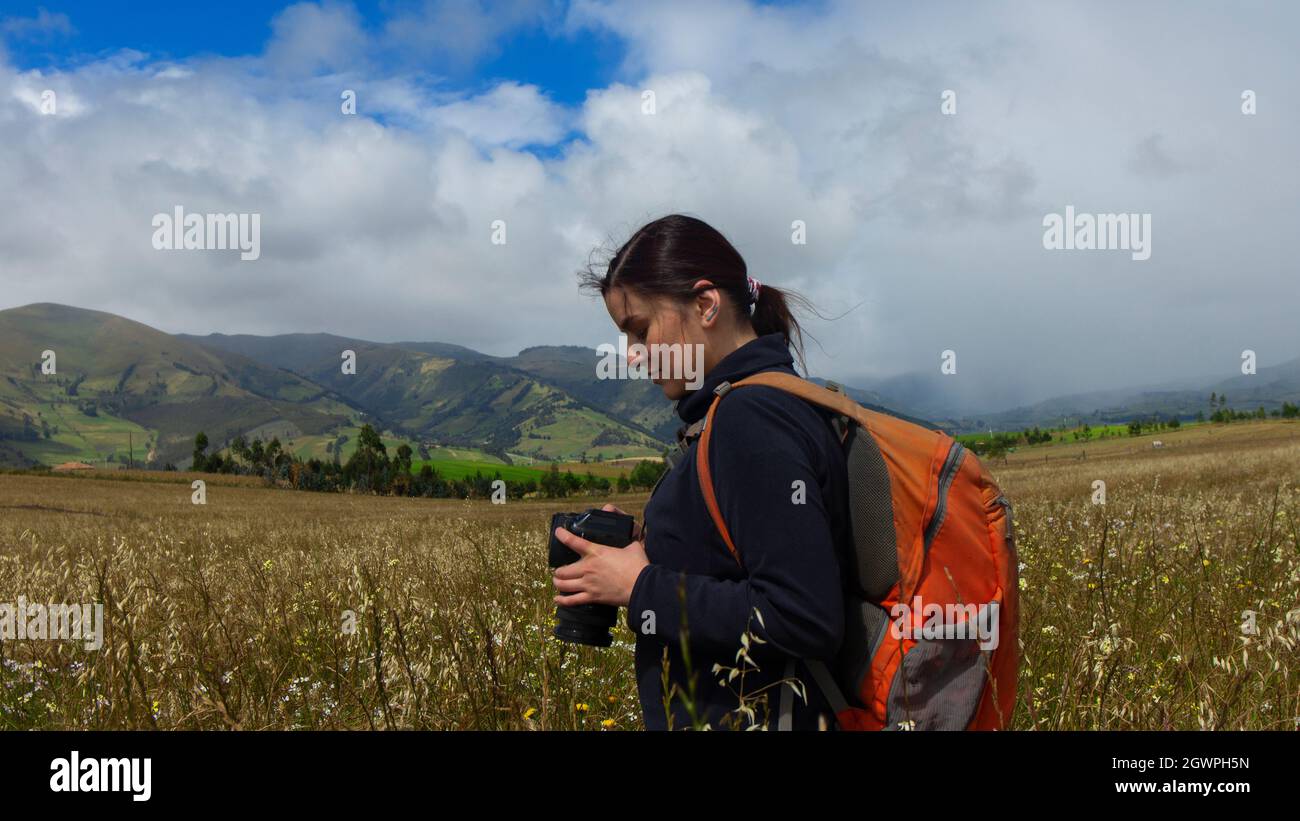 Wunderschöne hispanische Forscherin, von hinten gesehen, mit Rucksack, der Fotos auf ihrer Kamera an einem bewölkten Morgen inmitten eines gesät Feldes ansieht Stockfoto