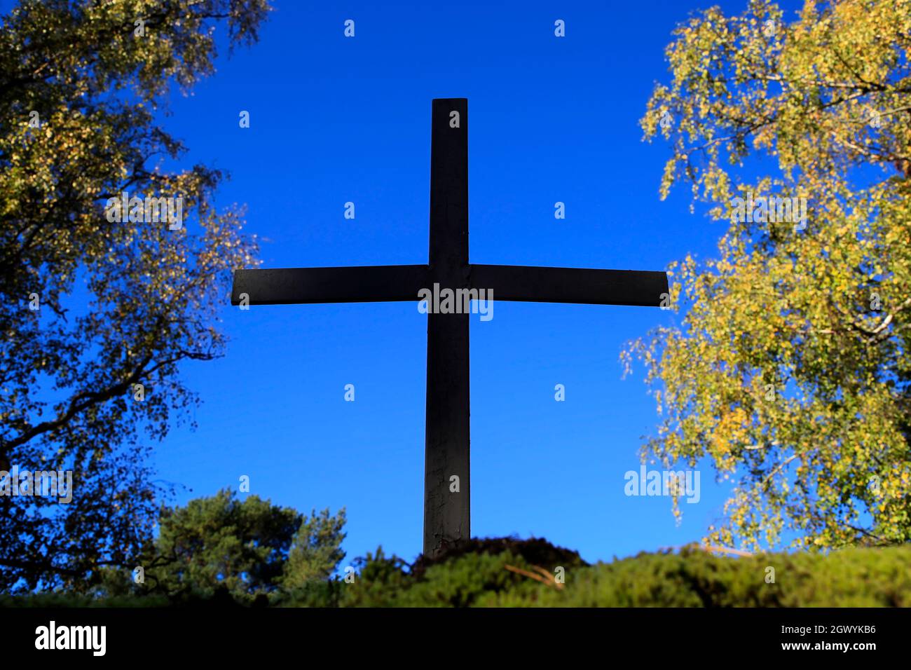 Metallisches Kreuz gegen blauen Himmel, mit herbstlichen Bäumen im Hintergrund. Stockfoto