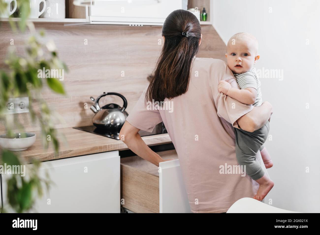 Beschäftigt Mutter Reinigung Küche mit Baby auf den Armen. Frau kocht und hält Säugling, müde Frau macht tägliche Hausaufgaben mit Kind. Stockfoto