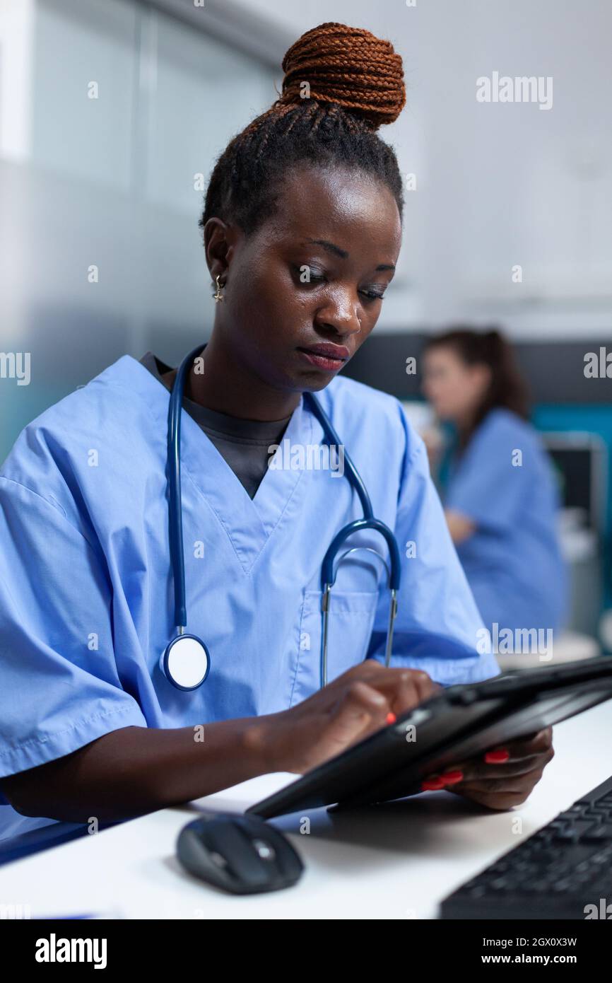 afroamerikanische Krankenschwester in medizinischer Uniform, die die Krankheitsverschreibung auf einem Tablet-Computer analysiert, der im Krankenhausbüro arbeitet. Arzt Astance Überwachung Krankheit schriftlich Gesundheitsversorgung Behandlung Stockfoto