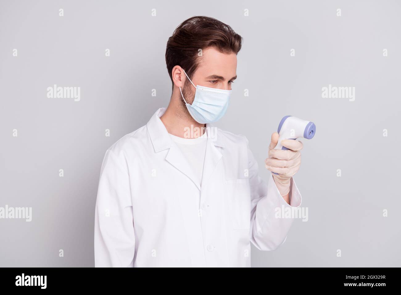 Foto von positiven jungen Doc Mann gekleidet weißen Mantel Messung Temperatur berührungslose Thermometer isoliert grauen Hintergrund Stockfoto