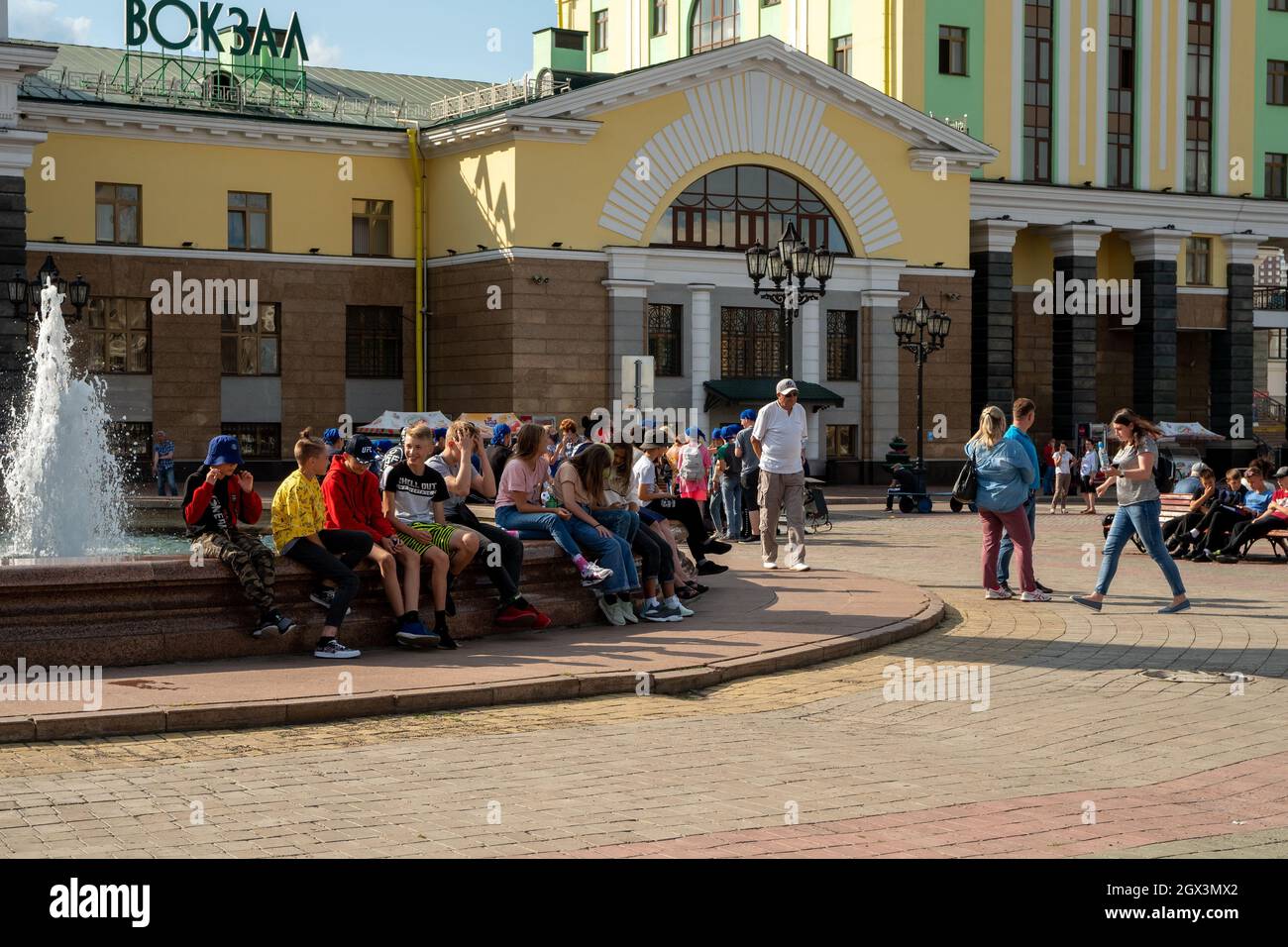 An einem sonnigen Sommertag sitzen Reisende am Brunnen auf dem Bahnhofsplatz vor der Kulisse des Hauptbahnhofs der Stadt. Stockfoto