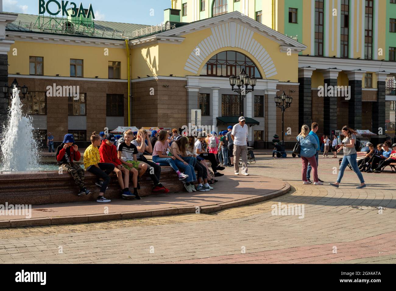 An einem sonnigen Sommertag sitzen Reisende am Brunnen auf dem Bahnhofsplatz vor der Kulisse des Hauptbahnhofs der Stadt. Stockfoto