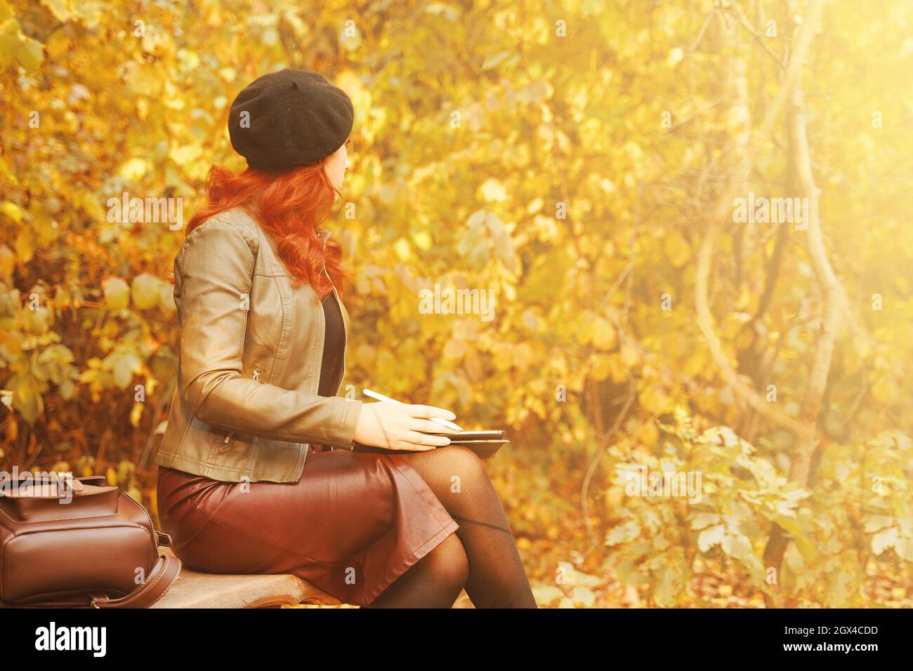 Herbstporträt. Hübsche Frau mit grafischem Tablet auf Parkbank. Rothaarige Mädchen in Baskenmütze und modische Jacke sieht weg. Lederrucksack auf einer Holzbank. Designer oder Freiberufler. Stockfoto