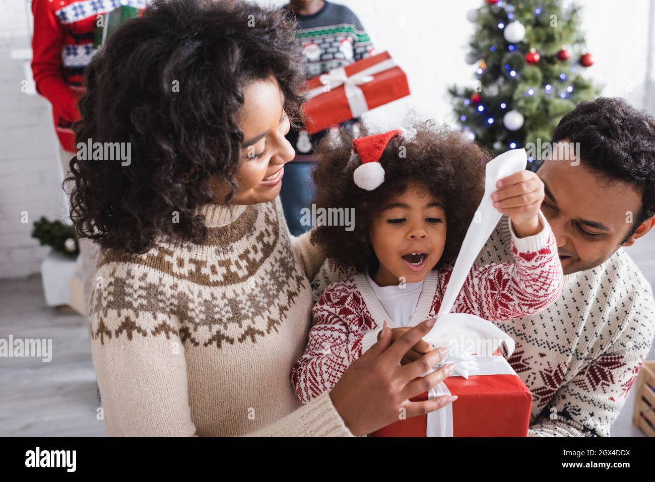 Erstaunt afroamerikanische Mädchen Eröffnung weihnachtsgeschenk in der Nähe glückliche Eltern und verschwommen weihnachtsbaum Stockfoto
