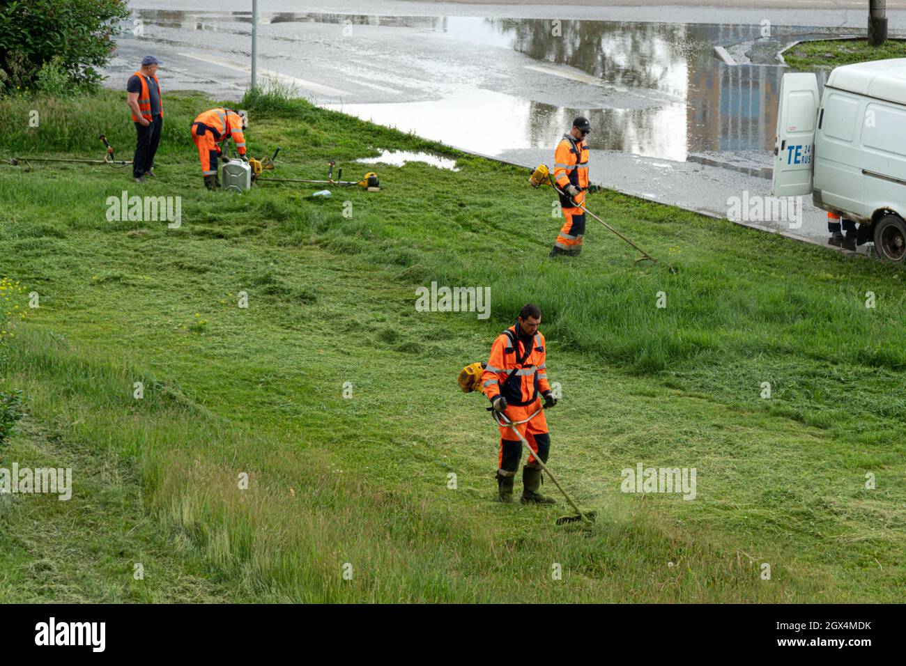 Eine Gruppe von Arbeitern in orangefarbenen Overalls mähen den Rasen an einem bewölkten Sommertag. Stockfoto