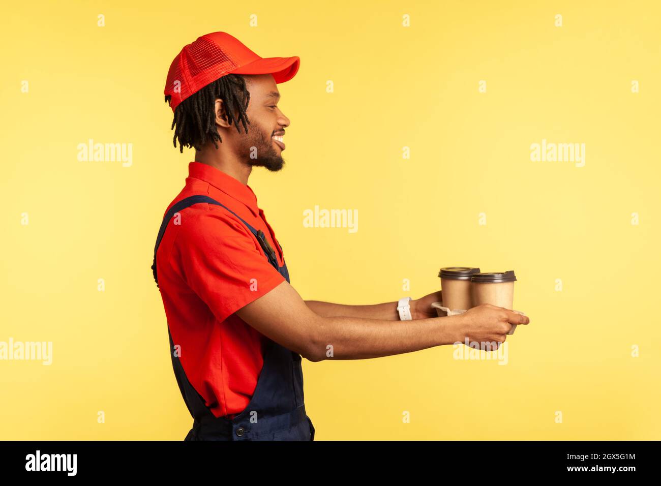 Lieferservice. Seitenansicht, freundlicher Lieferer in blauen Overalls, der Kaffee anbietet, Getränke in Einwegbechern gibt und lächelt. Innenaufnahme des Studios isoliert auf gelbem Hintergrund. Stockfoto