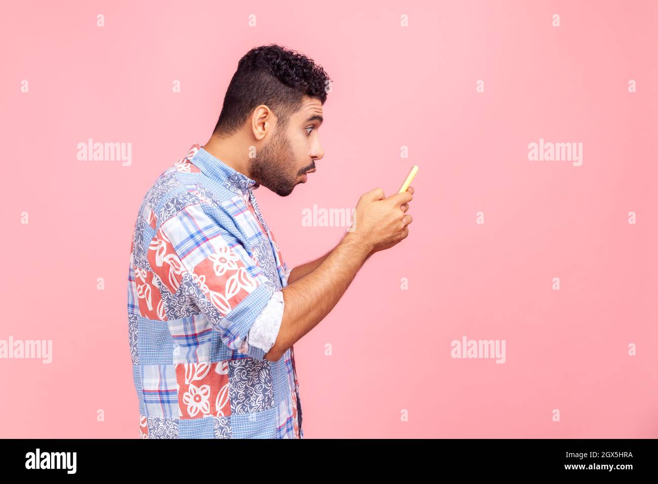 Seitenansicht Porträt von schockierten schönen Blogger jungen erwachsenen Mann mit Bart tragen in blauem Hemd mit Smartphone und beobachten Video mit staunenden Gesicht. Innenaufnahme des Studios isoliert auf rosa Hintergrund. Stockfoto
