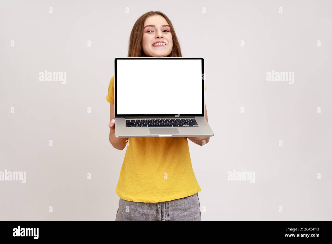 Porträt eines fröhlichen lächelnden, lockigen Teenagers im gelben Casual T-Shirt mit leerem Laptop-Bildschirm auf der Kamera, Bereich für Werbung. Innenaufnahme des Studios isoliert auf grauem Hintergrund. Stockfoto