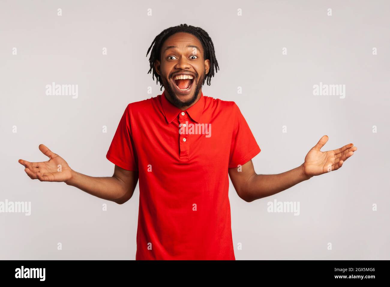 Toll. Attraktiver Mann mit Dreadlocks, der ein rotes T-Shirt im Casual-Stil trägt, überrascht ist, mit erhobenen Armen und offenem Mund in Staunen steht. Innenaufnahme des Studios isoliert auf grauem Hintergrund. Stockfoto