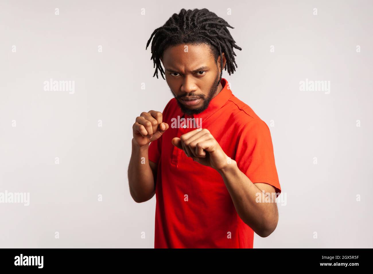 Aggressiver bärtiger Mann mit Dreadlocks, der ein rotes T-Shirt im lässigen Stil trägt, geballte Fäuste zum Boxen bereithält, Kampfkunsttrainer, Selbstverteidigung. Innenaufnahme des Studios isoliert auf grauem Hintergrund. Stockfoto